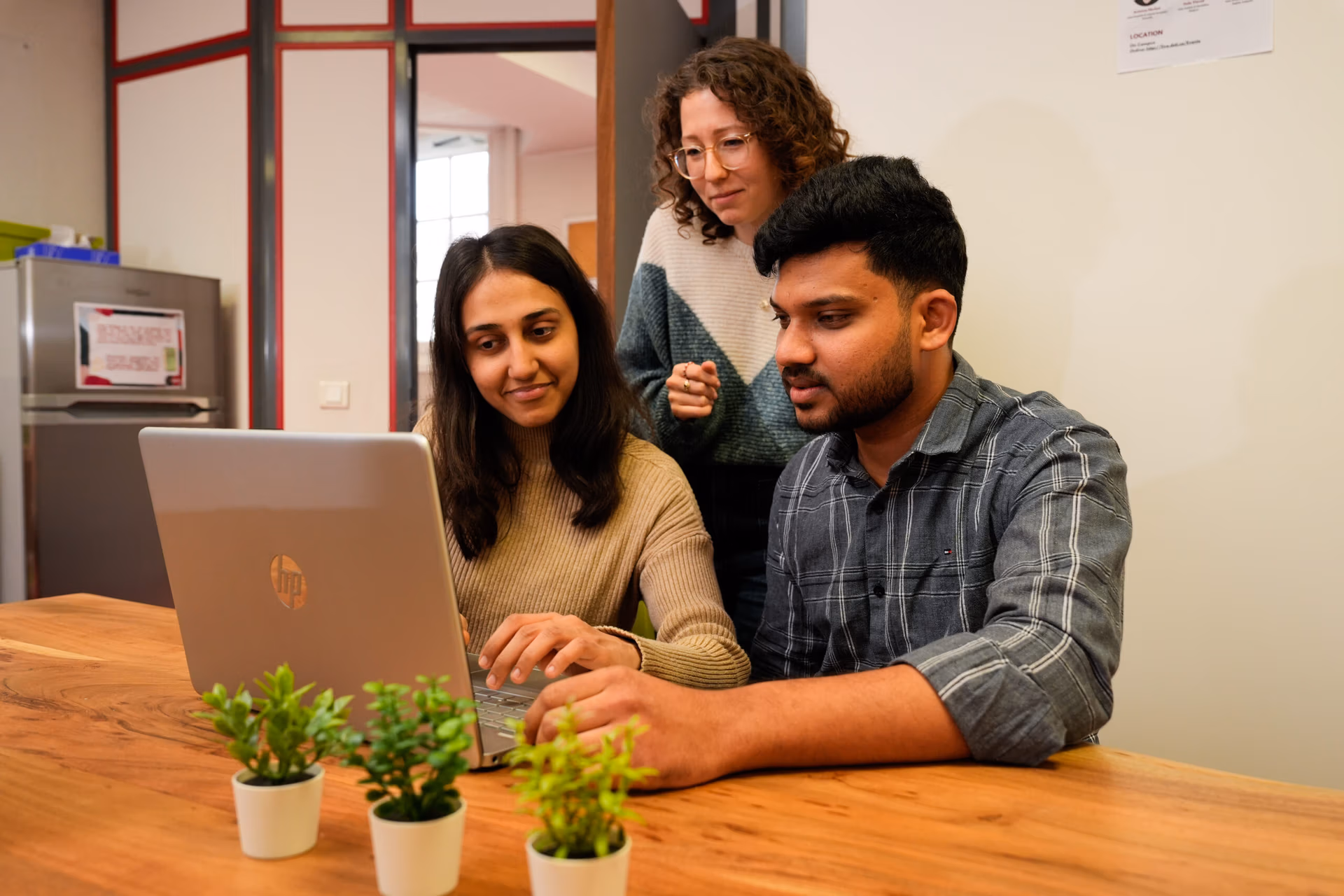 Three people talking in a casual indoor setting with a bulletin board and plant. Two of them are prospective students meeting with educational study abroad agents.